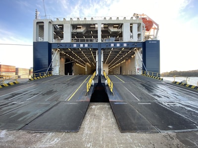 The image depicts the rear view of a large ferry or cargo ship with ramps lowered for vehicles to board. The interior cargo area is visible, well-lit with rows of lights along the ceiling. There are barriers and safety markings along the edges of the ramps, and a lifeboat is visible on the upper deck.