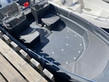 A small boat with two cushioned seats and clear windshields docked at a harbor. It has a sleek, dark-colored interior and visible hardware on the floor. There is a wooden dock beside the boat and parts of another boat are visible in the background.