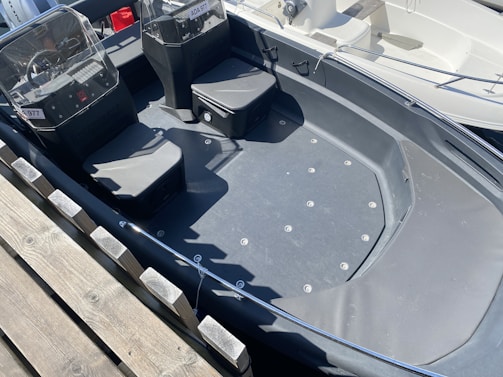 A small boat with two cushioned seats and clear windshields docked at a harbor. It has a sleek, dark-colored interior and visible hardware on the floor. There is a wooden dock beside the boat and parts of another boat are visible in the background.
