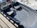 A small boat with two cushioned seats and clear windshields docked at a harbor. It has a sleek, dark-colored interior and visible hardware on the floor. There is a wooden dock beside the boat and parts of another boat are visible in the background.