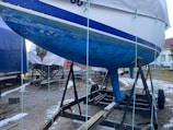 A boat with a blue hull is placed on a metal trailer in a yard. The hull shows signs of wear and algae growth. Other covered boats and a small building are in the background, with patches of snow on the ground.