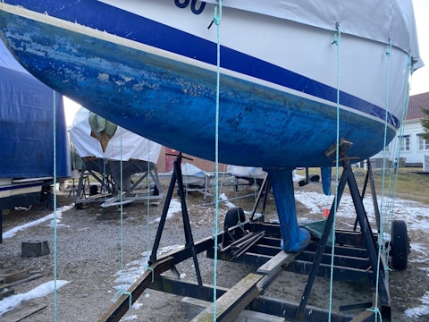 A boat with a blue hull is placed on a metal trailer in a yard. The hull shows signs of wear and algae growth. Other covered boats and a small building are in the background, with patches of snow on the ground.