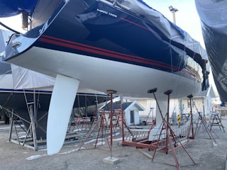 A large sailboat is mounted on stands in a boatyard. The hull is dark blue with red stripes, and it features a white keel. The boat is surrounded by a metal framework for support. The ground is gravel, and other boats and a small structure are visible in the background.