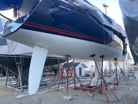 A large sailboat is mounted on stands in a boatyard. The hull is dark blue with red stripes, and it features a white keel. The boat is surrounded by a metal framework for support. The ground is gravel, and other boats and a small structure are visible in the background.