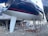 A large sailboat is mounted on stands in a boatyard. The hull is dark blue with red stripes, and it features a white keel. The boat is surrounded by a metal framework for support. The ground is gravel, and other boats and a small structure are visible in the background.
