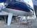 A large sailboat is mounted on stands in a boatyard. The hull is dark blue with red stripes, and it features a white keel. The boat is surrounded by a metal framework for support. The ground is gravel, and other boats and a small structure are visible in the background.