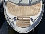 The bow of a boat is shown, featuring a wooden deck with neatly arranged planks. Two black ropes are tied to cleats at the front. The boat has metal railings along the sides and is moored, as indicated by the slack in the ropes. The water surrounding the boat is calm and dark.