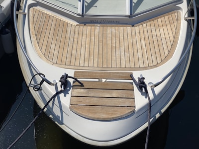 The bow of a boat is shown, featuring a wooden deck with neatly arranged planks. Two black ropes are tied to cleats at the front. The boat has metal railings along the sides and is moored, as indicated by the slack in the ropes. The water surrounding the boat is calm and dark.