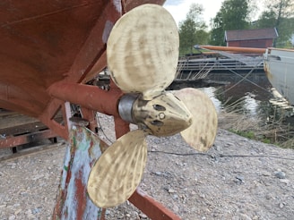 A large brass propeller attached to the underside of a boat. The area surrounding the propeller is made of reddish-brown metal. The setting appears to be outdoors near a body of water with nearby greenery and a wooden structure in the background.