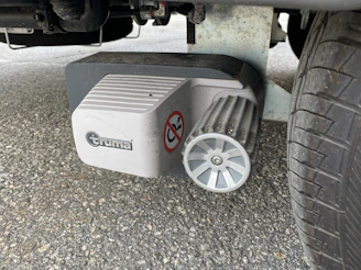 A mechanical component is attached to a tire, featuring a ribbed metallic wheel and a white plastic cover with a brand logo and a warning sticker. The ground is paved with asphalt.