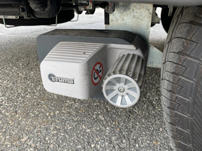 A mechanical component is attached to a tire, featuring a ribbed metallic wheel and a white plastic cover with a brand logo and a warning sticker. The ground is paved with asphalt.