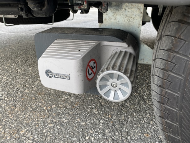 A mechanical component is attached to a tire, featuring a ribbed metallic wheel and a white plastic cover with a brand logo and a warning sticker. The ground is paved with asphalt.
