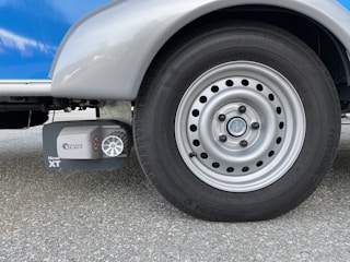 A close-up view of a silver vehicle wheel with a circular design featuring multiple holes around the rim. Adjacent to the wheel is a device labeled 'Truma Mover XT', which is likely a motorized mover for the vehicle. The surface is asphalt and the vehicle body is visible in shades of silver and blue.