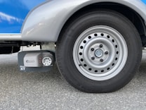 A close-up view of a silver vehicle wheel with a circular design featuring multiple holes around the rim. Adjacent to the wheel is a device labeled 'Truma Mover XT', which is likely a motorized mover for the vehicle. The surface is asphalt and the vehicle body is visible in shades of silver and blue.