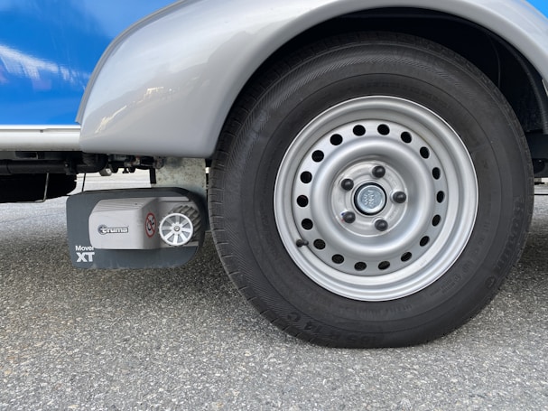 A close-up view of a silver vehicle wheel with a circular design featuring multiple holes around the rim. Adjacent to the wheel is a device labeled 'Truma Mover XT', which is likely a motorized mover for the vehicle. The surface is asphalt and the vehicle body is visible in shades of silver and blue.