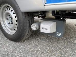 A close-up view of a vehicle wheel on a textured asphalt surface, featuring a Truma Mover XT device attached near the tire. The tire tread and the mechanical components of the mover stand out prominently.