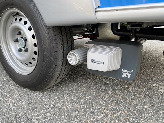 A close-up view of a vehicle wheel on a textured asphalt surface, featuring a Truma Mover XT device attached near the tire. The tire tread and the mechanical components of the mover stand out prominently.