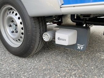 A close-up view of a vehicle wheel on a textured asphalt surface, featuring a Truma Mover XT device attached near the tire. The tire tread and the mechanical components of the mover stand out prominently.