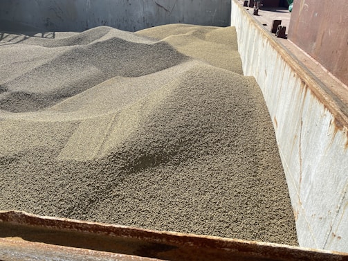 A large quantity of granular material, likely grain, is piled in a metal container or storage bin. The surface of the material forms gentle, undulating mounds, suggesting it has been recently poured. The container shows signs of rust and wear.