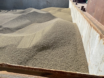 A large quantity of granular material, likely grain, is piled in a metal container or storage bin. The surface of the material forms gentle, undulating mounds, suggesting it has been recently poured. The container shows signs of rust and wear.