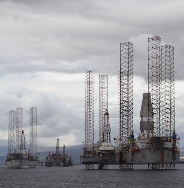 Several large offshore oil drilling platforms with tall lattice towers standing in the sea. The platforms are surrounded by a slightly cloudy sky.