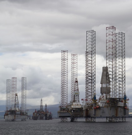 Several large offshore oil drilling platforms with tall lattice towers standing in the sea. The platforms are surrounded by a slightly cloudy sky.