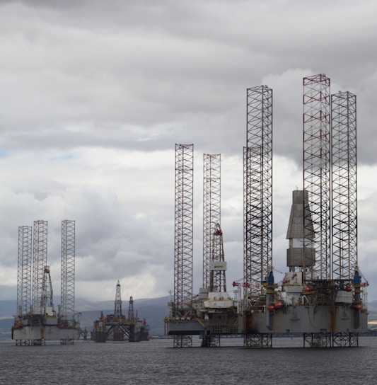 Several large offshore oil drilling platforms with tall lattice towers standing in the sea. The platforms are surrounded by a slightly cloudy sky.