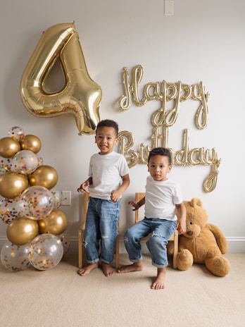 Two young children are sitting on wooden chairs next to a large brown teddy bear. They are surrounded by gold and transparent balloons with colorful confetti inside. Gold balloons spell out 'Happy Birthday' with a large gold number 4 balloon nearby. The children are dressed in casual white t-shirts and jeans, and they appear to be celebrating a birthday.