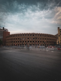 A large, historic-looking arena with multiple arches lines the upper levels, surrounded by an urban setting with tall buildings. The structure has a classic architectural style with repetitive arched windows. In front of the arena, there is a street visible with traffic lights and a group of bicycles parked.