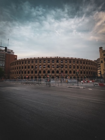 A large, historic-looking arena with multiple arches lines the upper levels, surrounded by an urban setting with tall buildings. The structure has a classic architectural style with repetitive arched windows. In front of the arena, there is a street visible with traffic lights and a group of bicycles parked.