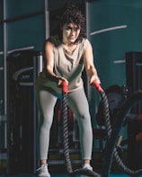A woman is in a gym holding battle ropes with both hands, appearing focused and engaged in an exercise. She wears a light-colored tank top, leggings, and athletic shoes. The background includes gym equipment, and the atmosphere suggests a fitness environment.