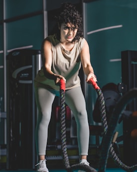 A woman is in a gym holding battle ropes with both hands, appearing focused and engaged in an exercise. She wears a light-colored tank top, leggings, and athletic shoes. The background includes gym equipment, and the atmosphere suggests a fitness environment.