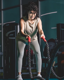 A woman is in a gym holding battle ropes with both hands, appearing focused and engaged in an exercise. She wears a light-colored tank top, leggings, and athletic shoes. The background includes gym equipment, and the atmosphere suggests a fitness environment.