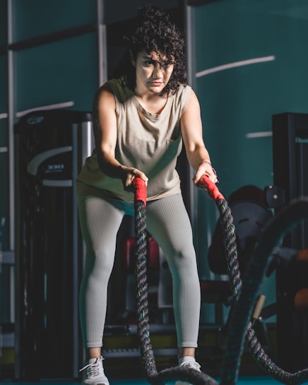 A woman is in a gym holding battle ropes with both hands, appearing focused and engaged in an exercise. She wears a light-colored tank top, leggings, and athletic shoes. The background includes gym equipment, and the atmosphere suggests a fitness environment.
