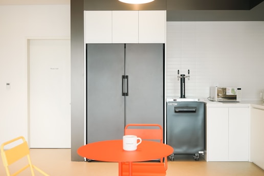 A modern kitchenette features a large gray refrigerator flanked by white cabinetry. A compact bar unit with a dual tap system sits beside a microwave on the countertop. A bright orange circular table with a matching orange chair and a yellow chair are positioned in the foreground, with a white mug resting on the table. The room is well-lit with a clean, minimalist style, and there is a door labeled 'Music Hall' on one side.