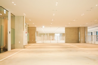 A spacious, well-lit interior of an empty room with a minimalistic design. The floor and walls are primarily beige and off-white, with a large ceiling featuring numerous small recessed lights. There are glass doors on the left side, and the end of the room is sectioned with a metal railing that divides the space.