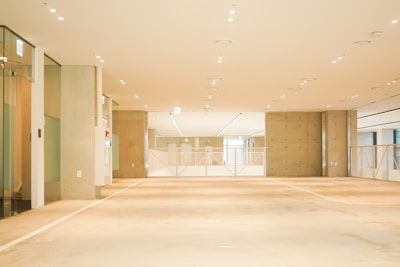 A spacious, well-lit interior of an empty room with a minimalistic design. The floor and walls are primarily beige and off-white, with a large ceiling featuring numerous small recessed lights. There are glass doors on the left side, and the end of the room is sectioned with a metal railing that divides the space.