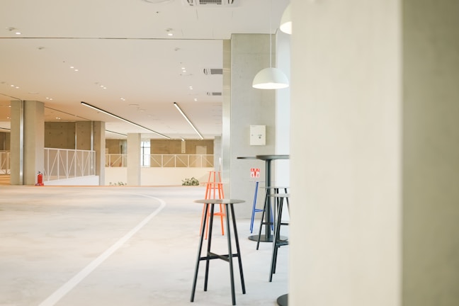 A spacious and well-lit interior with modern minimalist design. The floor has a smooth grey finish, while the ceiling features a series of recessed lights and linear light fixtures. In the foreground, there are a few high tables accompanied by tall stools in black, orange, and blue colors. Large rectangular columns divide the space, and a fire extinguisher is mounted on one of them. Pendant lights hang from the ceiling, providing focused illumination. The background features railing partitions and windows that allow natural light to enter.