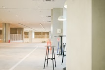 A spacious and well-lit interior with modern minimalist design. The floor has a smooth grey finish, while the ceiling features a series of recessed lights and linear light fixtures. In the foreground, there are a few high tables accompanied by tall stools in black, orange, and blue colors. Large rectangular columns divide the space, and a fire extinguisher is mounted on one of them. Pendant lights hang from the ceiling, providing focused illumination. The background features railing partitions and windows that allow natural light to enter.