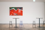 A minimalist interior space with a colorful painting on a white wall. The painting features abstract figures and vivid colors such as red, green, and white. Below the painting are modern, round tables with mismatched stools, including a distinct blue stool.