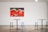 A minimalist interior space with a colorful painting on a white wall. The painting features abstract figures and vivid colors such as red, green, and white. Below the painting are modern, round tables with mismatched stools, including a distinct blue stool.