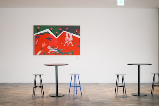 A minimalist interior space with a colorful painting on a white wall. The painting features abstract figures and vivid colors such as red, green, and white. Below the painting are modern, round tables with mismatched stools, including a distinct blue stool.