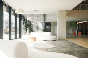 A spacious, modern interior with large floor-to-ceiling windows allowing natural light to fill the space. The area features white, minimalist furniture, including sofas and a table, complemented by potted green plants. The floor is a mix of gray tiles and wood. Above, there are sleek white pendant lights. A few orange traffic cones and signs are visible in the background.