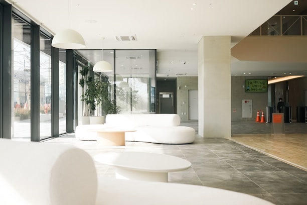 A spacious, modern interior with large floor-to-ceiling windows allowing natural light to fill the space. The area features white, minimalist furniture, including sofas and a table, complemented by potted green plants. The floor is a mix of gray tiles and wood. Above, there are sleek white pendant lights. A few orange traffic cones and signs are visible in the background.