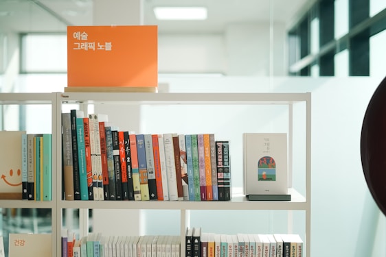A bookshelf filled with a variety of books in different colors and sizes. There is an orange sign with Korean text on top of the shelf. The books are organized neatly, and the shelf is located in a well-lit room with large windows in the background.