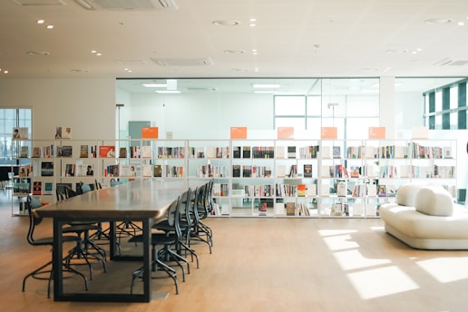 A spacious, modern library interior featuring a long wooden table with several chairs lined up and bookshelves filled with various books. Large windows allow natural light to flood the space, casting shadows on the wooden floor. The atmosphere is minimalist and organized with a comfortable white sofa in the foreground.