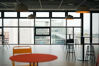 An indoor space features several round tables and bar stools with pendant lights hanging from the ceiling. Large windows offer a view of modern buildings outside, allowing natural light to fill the room.
