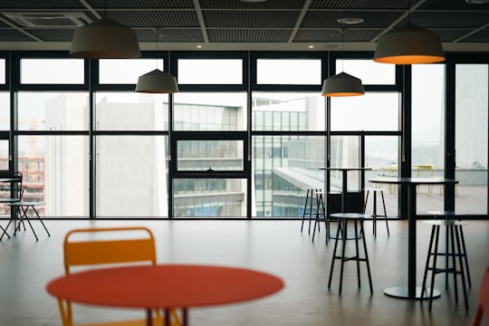 An indoor space features several round tables and bar stools with pendant lights hanging from the ceiling. Large windows offer a view of modern buildings outside, allowing natural light to fill the room.