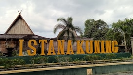 Large yellow letters spelling 'ISTANA KUNING' are prominently displayed in front of traditional wooden buildings with steeply pitched roofs. Tall palm trees and lush greenery surround the area under a slightly overcast sky.