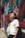 A person with headphones around their neck leans against a graffiti-covered brick wall in an alley. They are holding a white bag and wearing a white T-shirt. The scene is brightly lit with colorful urban art surrounding them.
